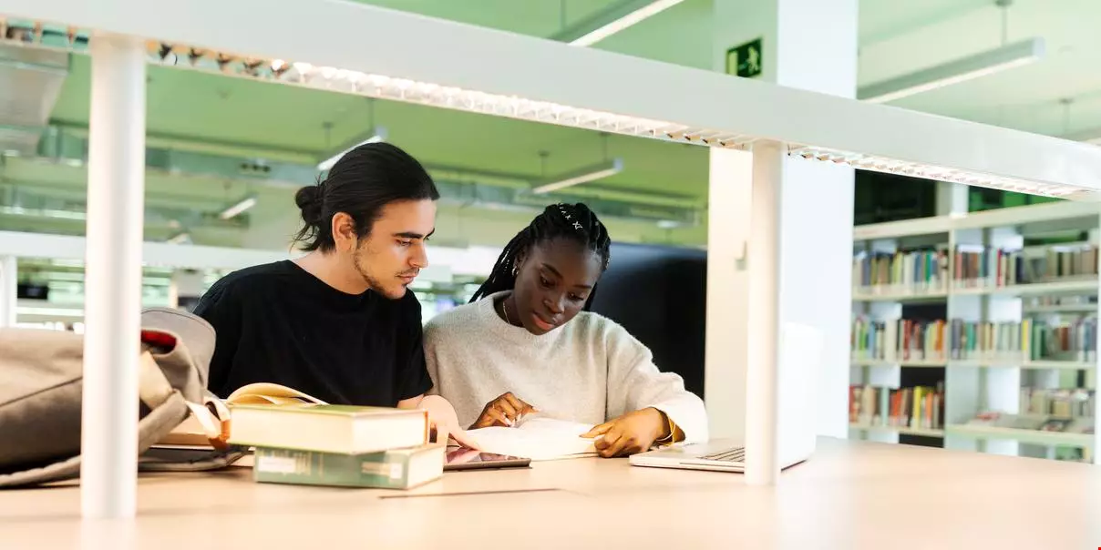 En manlig och kvinnlig student sitter tätt ihop och läser i en bok tillsammans  i ett bibliotek.